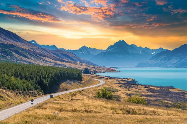 A road running beside a turquoise lake toward snowy mountains